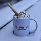 Mug of hot chocolate with whipped cream and a cinnamon stick on a snowy surface, featuring 'The Coffee Lab' branding.